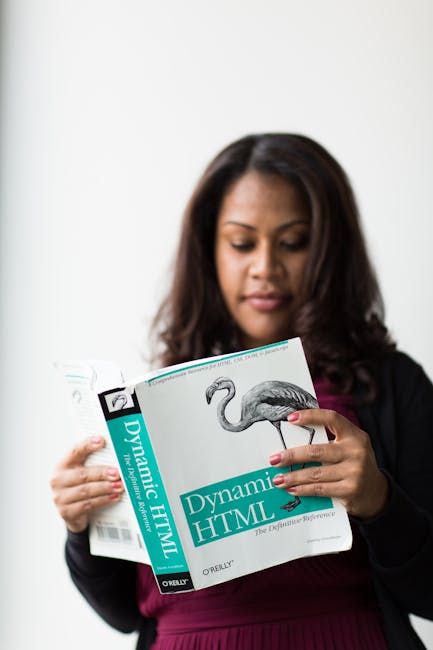 The image features a woman with dark hair, dressed in a black blazer and a dark pink top, standing indoors against a plain, light-colored background. She is holding a book titled 'Dynamic HTML' by O'R
