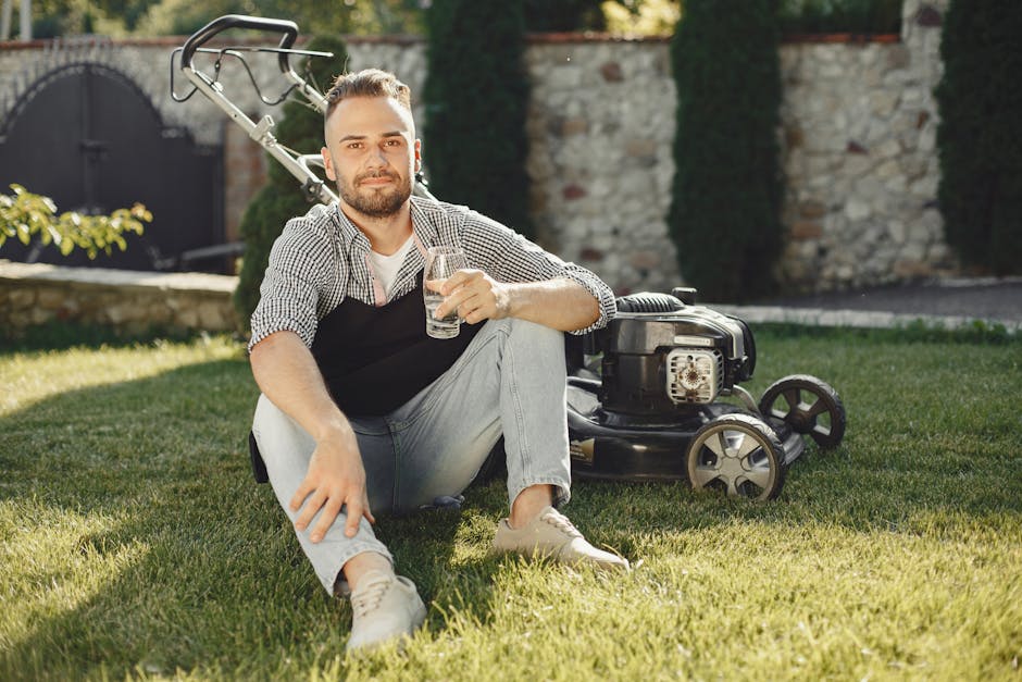 A man with a beard and short hair, dressed in a checkered shirt and light-colored jeans, is sitting on a well-maintained grassy lawn in a garden, holding a glass of water and smiling at the camera. Be